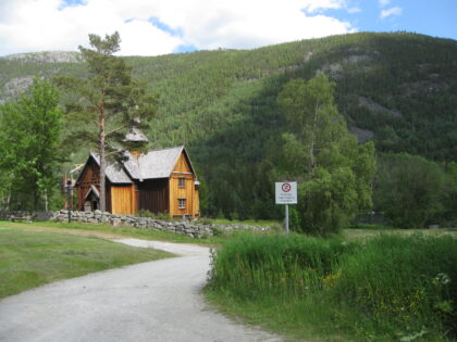i enden av en svingete vet stpr en liten, brunoransje stavkirke. Det er sommer og landskapet er grønt. Veien er grå. Foran til høyre for stavkirken står det et veiskilt.