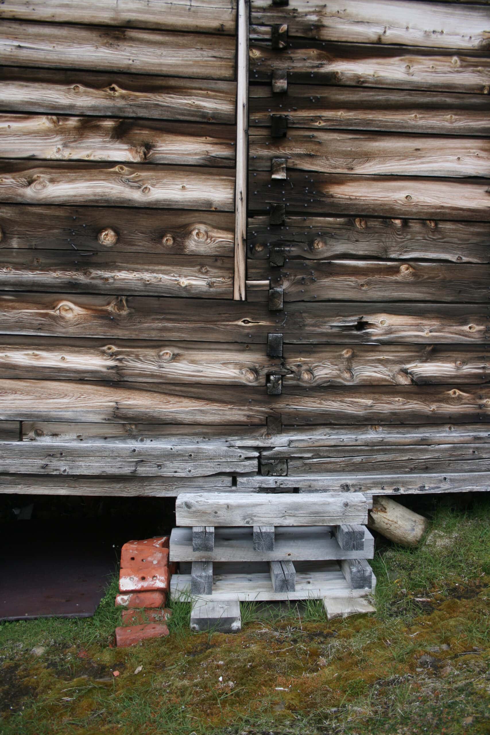 Foto som viser hvordan jordsig har dyttet den gamle pælen over ende slik at bygningen har seget nedover. Sysselmannen har tidligere satt nye, midlertidige pakke-fundamenter under bygget som erstatning for de gamle pælene.