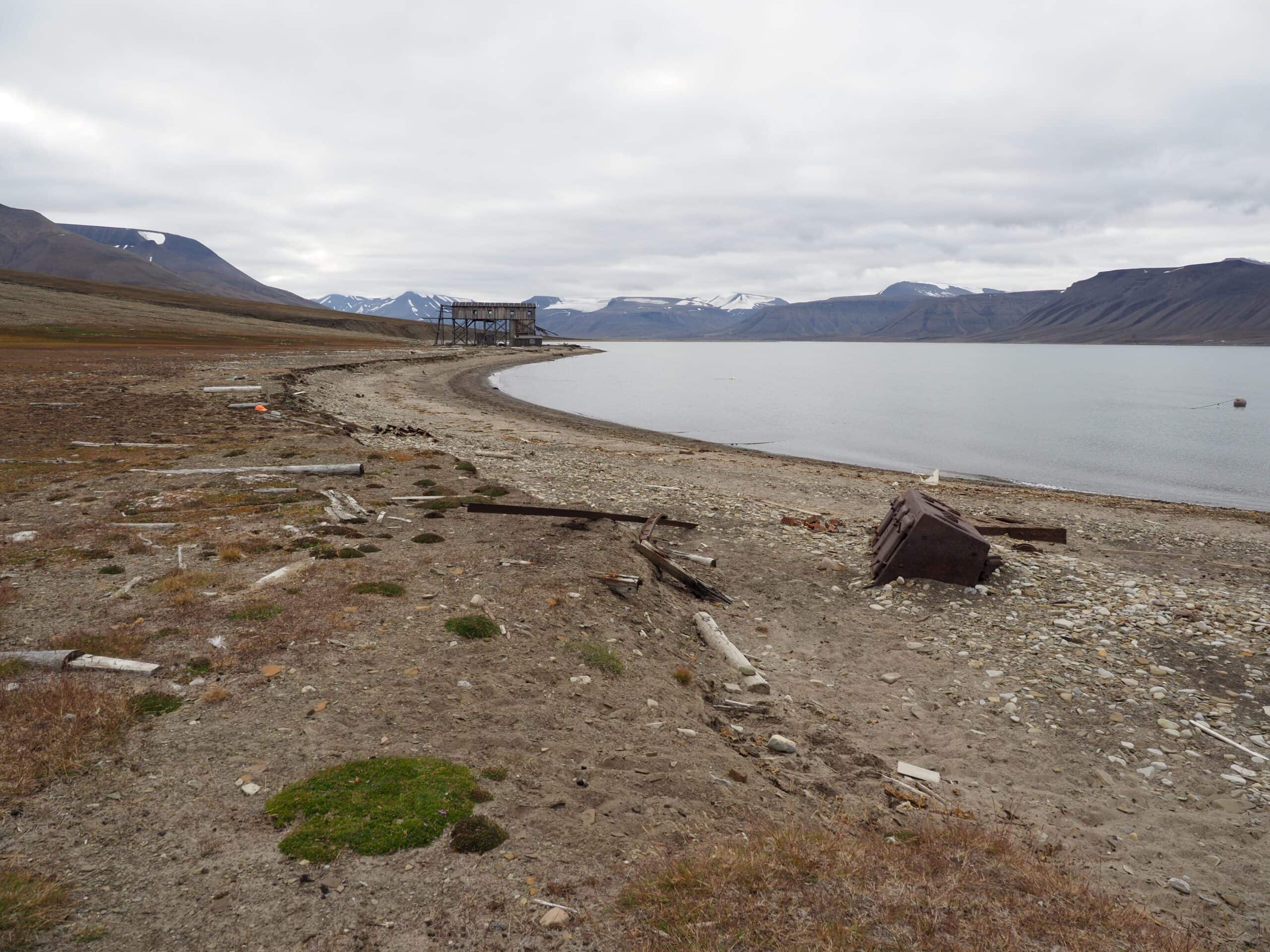 Bølger og av og til også fjordis – selv om det er svært lenge siden isen la seg på Adventfjorden nå – bryter ned strandbrinken på Hiorthhamn. Restene etter et lokomotiv og dens skinnegang ligger nedenfor erosjonskanten og vitner om sterke krefter – selv om selve kanten bare er noen 10-talls cm høy. Kysterosjon er en av nebrytningsmekanismene som ANH-prosjektet ser nærmere på i Hiorthhamn. Foto: AC Flyen/NIKU