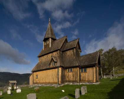 Bildeav Urnes stavkirke. Kirken er bygget av tre og har en oransjeaktig brunfarge. Det er sommer, grønt gress og blå himmel med noen skyer. Vi ser noen gravsteiner i forgrunnen, og noen trær i høyre billedkant.