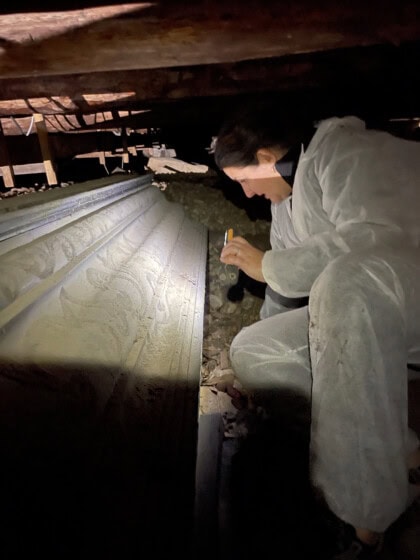 Hanne Ekstrøm Jordahl undersøker kiste under gulvet i Solum kirke, Hun er iført en hvit heldekkende drakt og hodelykt. Rommet er mørkt og trangt.