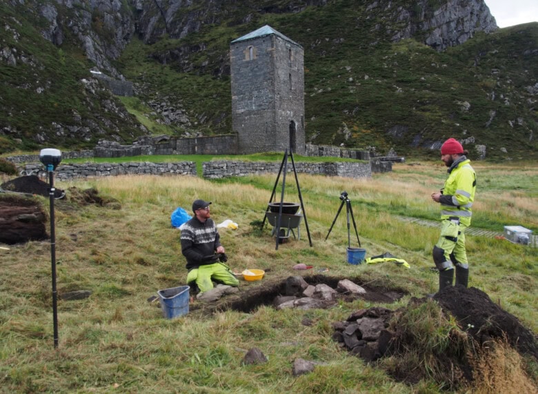 To arkeologer graver i bakken foran ruinen av Selja kloster- og helgenanlegg.