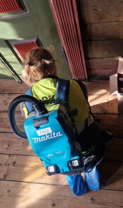 Konservator sitter på kne med en blå støvsuger på ryggen. HUn er iført en gul vest med NIKU-logo, og befinner seg inne i Finnegården på Bryggen i Bergen.