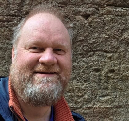 Selvportrett av en skjeggete mann foran en bergvegg. På bergveggen er det risset inn et ansikt, en helleristning.