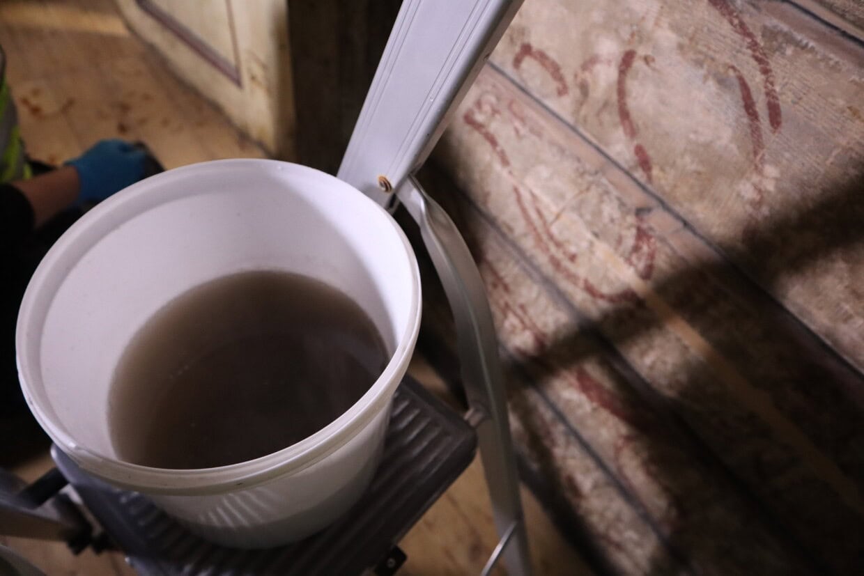 En hvit vaskebøtte med skittent vann på en stige. Stigen står ved siden av en laftet vegg i Finnegården på Bryggen i Bergen.