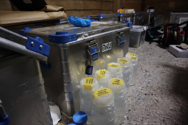 En stårlkoffert med NIKU-logo står i et rom med laftede vegger. Den inneholder utstyr som konservatorer bruker for å rense overflatene i Finnegården, Bryggen i Bergen. Foran kofferten står det gjennomsiktige flasker med blankt innhold. Innholdet står beskrevet på gul tape på lokket av flaskene. 