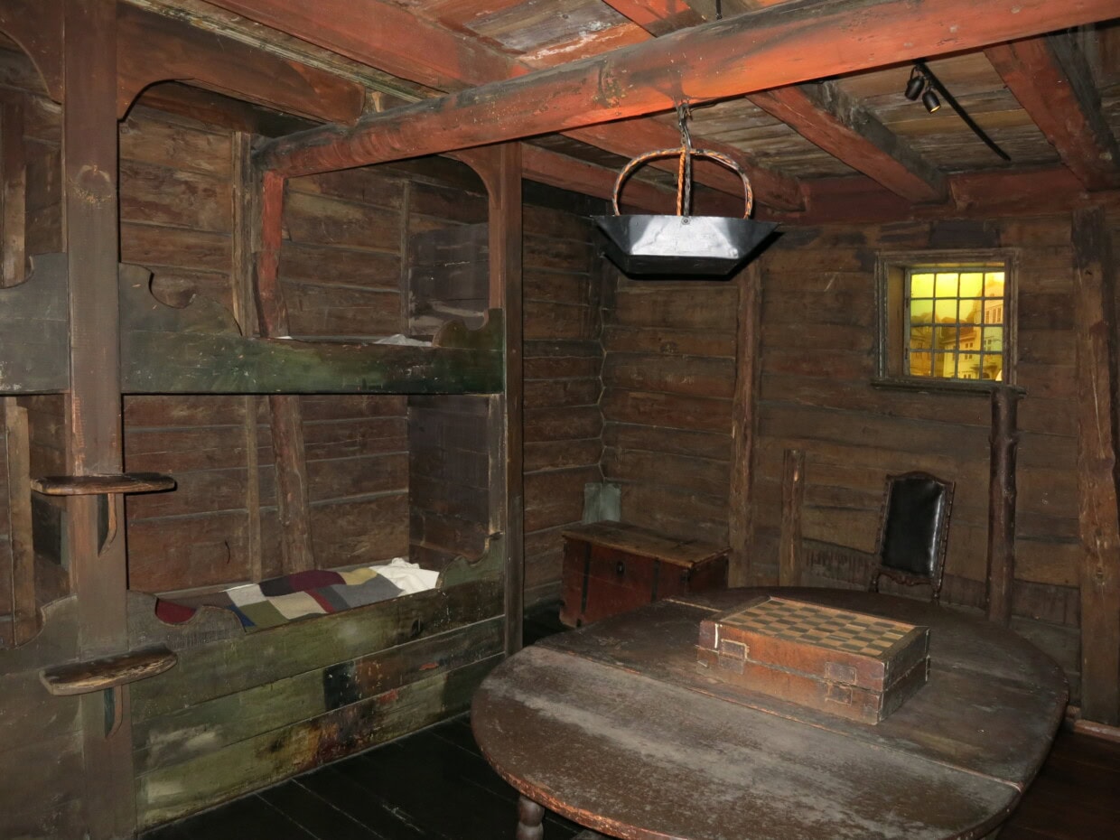 Bilde av et rom fra det Hanseatiske museum vwed bryggen i Bergen. Rommet er innredet som et hanseatisk bryggehus, med fire greønne veggfaste senger til venstre i rommet ig en kirste i hjørnet. På et ovalt bord står et sjakkbrett. Veggene er av tømmer og bjelkene i taket er malt røde.