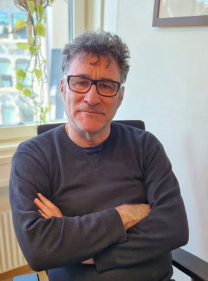 Portrett av Michael Derrick. Han sitter på en kontorstol med armene i kors, iført en mørk blå genser og briller. 