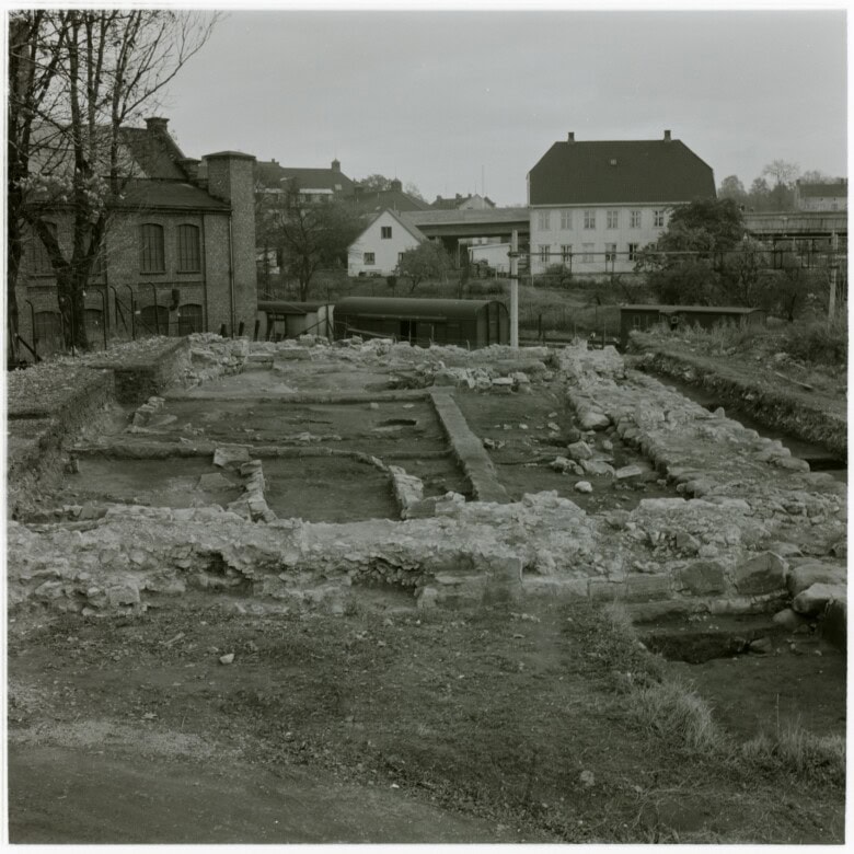 Svart-hvitt fotografi av ruinene etter kongsgården fra utgravning på 1960-tallet. I bakgrunnen ses mer moderne bebyggelse, blant annet Saxegården og Lokomotivverkstedet.