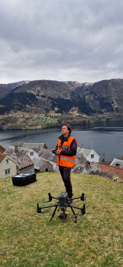 En mann med svarte klær og oransje vest står foran en husklynge i bratt vestlandslandskap, Han har en stor drone foran seg, som han forbereder seg på å fly.