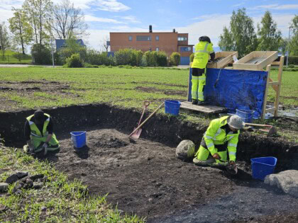 Tre arkeologer jobber på et lite område som er åpnet ute på et jorde. To arkeologer sitter på knærne nede i utgravningsfeltet, mens den andre arkeologen sålder masser ved en såldestasjon i bakgrunnen