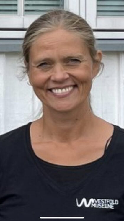 Portrett av en smilende kvinne med lyst hår i hestehale. Hun har på seg en svart t-skjorte med Vestfold museene -logo, og står foran en hvit trebygning.