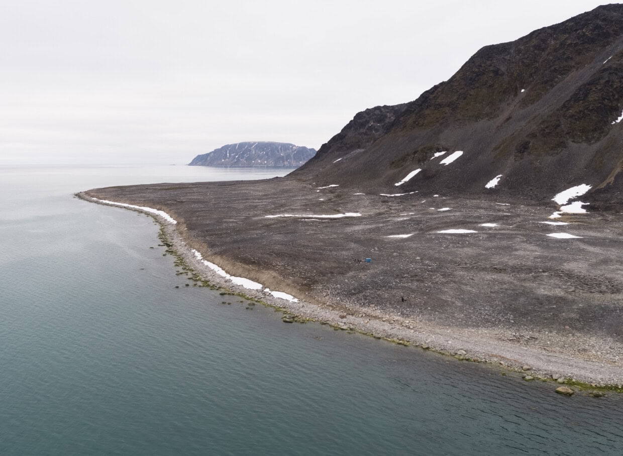 Vi ser en kyststrekning mot havet med fjell i høyden, og en øy i bakgrunnen. Det er steinete og spor av snø.