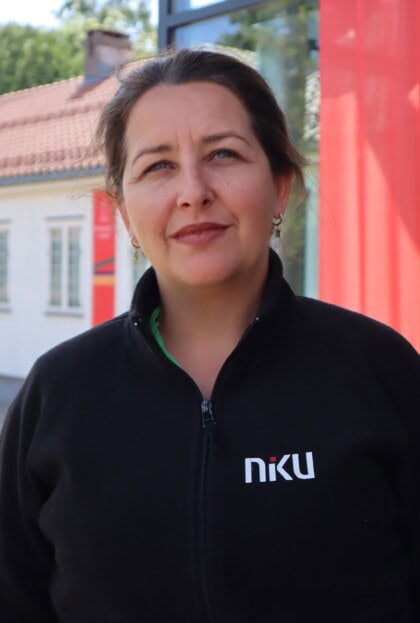 En kvinne som smiler forsiktig mens hun ser inn i kamera. hun har mørkt hår i hestehale, og en svart jakke med NIKU-logo på. Hun står foran en hvit trebygning.