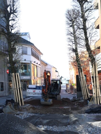 En gravemaskin som graver i en brolagt gate i Tønsberg. Det er trehus på den ene siden av gata og en mer moderne bygning på den andre. Graveområdet er sperret av med byggegjerder.