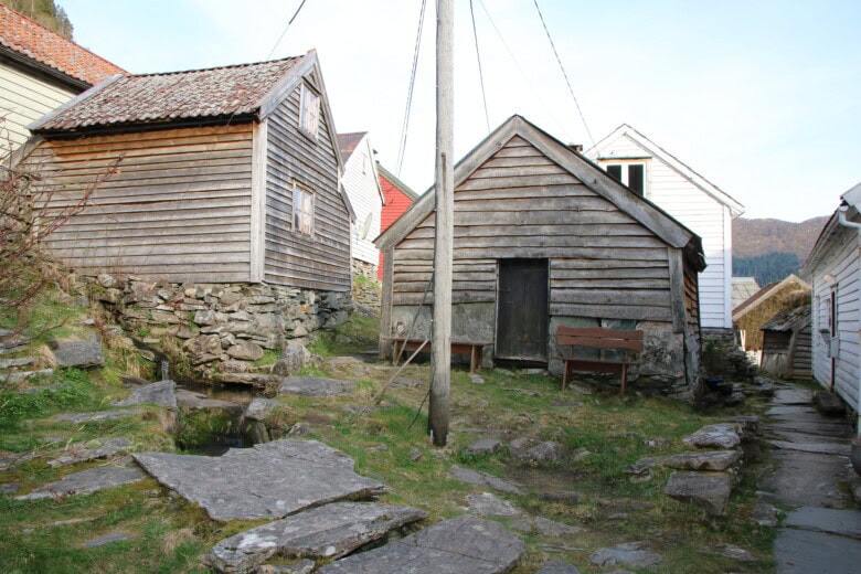 Bilde av to trebygninger tett i tett på Havrå. Til venstre i bildet renner en bekk med heller delvis plassert over, og til høyre er en rekke hus plassert langs en trang, hellelagt gate. Midt i bildet står en lyktestolpe, og to benker er plassert foran bygningen midt i bildet.
