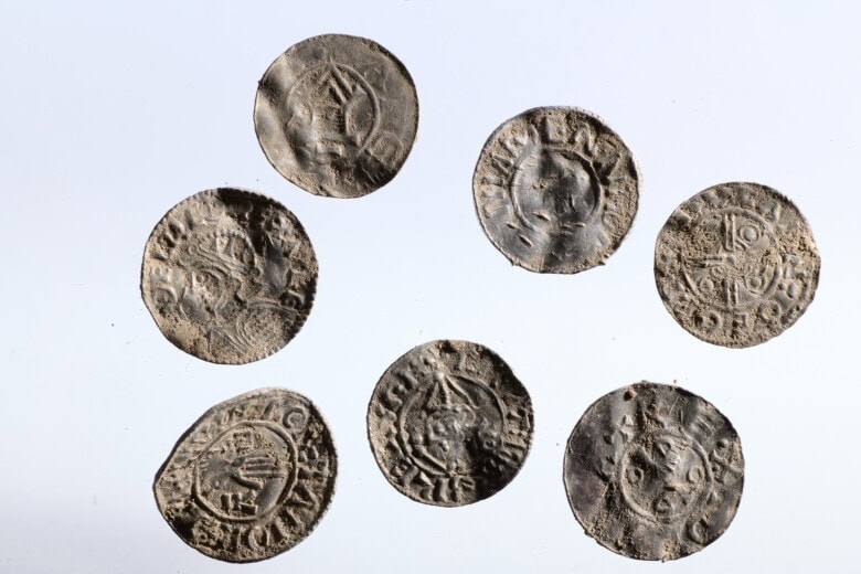 Syv mynter fra overgangen mellom vikingtid og tidlig middelalder. De er del av det store myntfunnet på Mørstad, Innlandet. Myntene ligger på hvit bakgrunn.