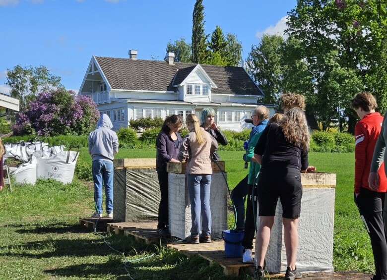 Flere unge mennesker står med ryggen til kamera og sålder masser på en arkeologiesk utgravning. Det er fint vær, sommer og i bakgrunnen av bildet står det et stort, hvitt hus.