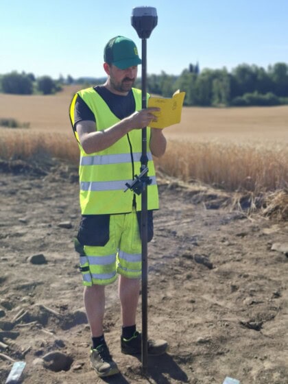 En mannlig arkeolog iført gult vernetøy måler inn graver på Valum utenfor Hamar. Han noterer i en gul bok.